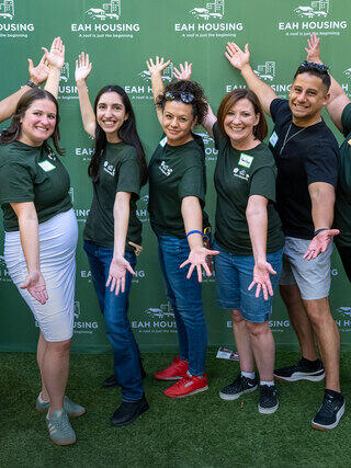 A group of five people in matching shirts pose cheerfully with outstretched arms against a green backdrop featuring "EAH Housing."