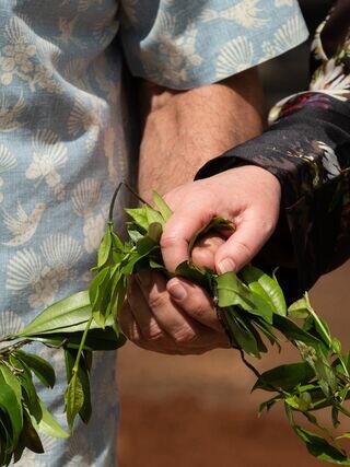 Two hands grasp a bundle of green leaves, symbolizing unity, connection, and nature. Soft fabrics in the background add a warm touch.