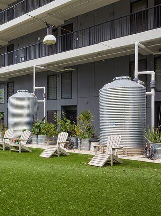 Two large, silver storage tanks stand next to white lounge chairs on a grassy area, surrounded by plants and a modern building façade.