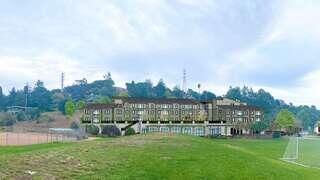 A large, multi-story building with a modern design sits at the edge of a green field, surrounded by hills and trees under a cloudy sky.