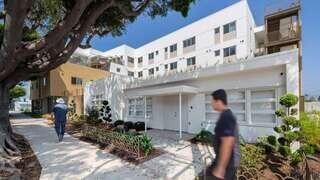 A modern multi-story building with a clean white facade, surrounded by landscaped greenery, and two pedestrians walking along the path.