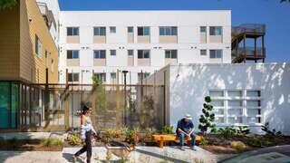A modern, multi-story building features a mix of white and beige exteriors, large windows, and landscaping with trees and greenery.