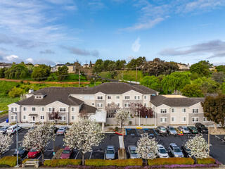 A two-story residential building surrounded by blooming trees and greenery, with a parking lot filled with cars underneath a blue sky.