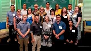 A diverse group of individuals stands together, smiling for a photo at an event, with a green backdrop displaying the organization's logo.