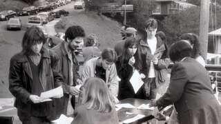 A group gathers around a table outdoors, engaged in discussion and exchanging papers, while parked cars line a hill in the background.