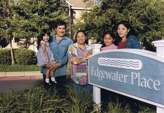 A family stands together outside a residential sign, smiling and posing amidst lush greenery. The atmosphere feels warm and welcoming.