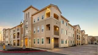 A modern multi-story apartment complex with a light beige exterior, featuring balconies and large windows, set in a spacious environment.
