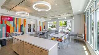A modern, brightly lit space features a colorful accent wall, sleek furniture, and a spacious kitchenette, creating a vibrant communal area.