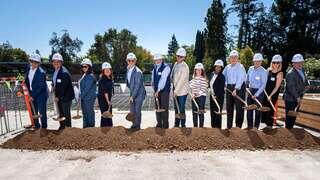 A group of individuals in hard hats gathers to participate in a groundbreaking ceremony, holding shovels over a mound of dirt.