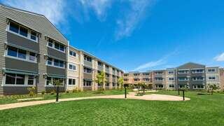 Exterior view of Aloha Iā Halewilikō, featuring a multi-story residential building with neutral tones, landscaped green space, and walking paths under a bright blue sky.
