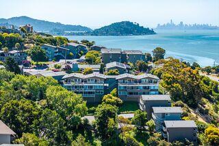 A scenic coastal view featuring lush greenery and contemporary buildings, with hills and the distant skyline of a city across the bay.