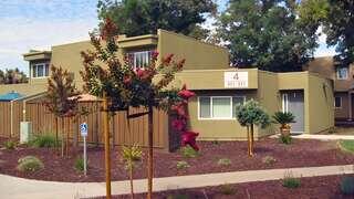 Don De Dios Apartments, EAH Housing, exterior featuring a single-story residential building with a sloped roof, private entryways, and landscaped front gardens with flowering plants and trimmed trees.