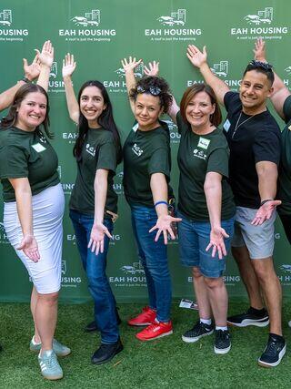 A group of cheerful individuals in matching green shirts, posing with outstretched arms in front of a branded backdrop.