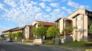 A row of modern residential buildings features landscaped greenery and a clear blue sky with clouds, creating a welcoming atmosphere.