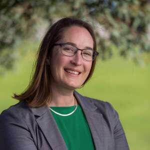 A woman with glasses smiles confidently, wearing a green top and grey blazer, with greenery in the background.