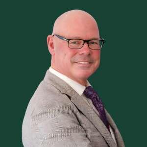 A smiling, bald man in a gray suit and purple tie poses confidently against a solid green background. Glasses accentuate his features.