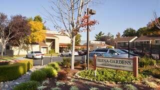 Elena Gardens Apartments, EAH Housing, exterior featuring a landscaped community entrance with signage, parking area, and surrounding residential buildings with trees and greenery.