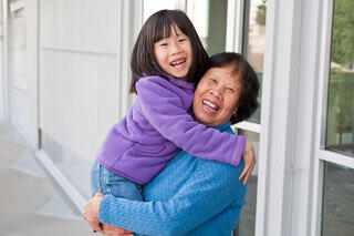A joyful grandmother holds her smiling granddaughter, both sharing a moment of love and happiness in a bright, outdoor setting.