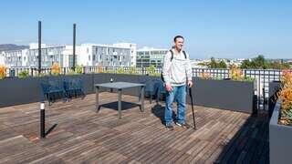 A young man stands on a rooftop terrace with wooden flooring, surrounded by plants and modern buildings under a clear blue sky.