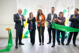 A group of six people, including a woman in an orange blazer, celebrate a ribbon-cutting ceremony with balloons and a green ribbon.