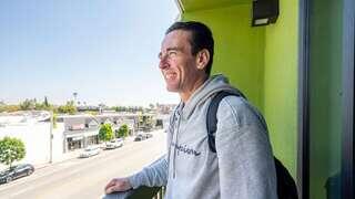 A man smiles while leaning on a balcony railing, enjoying a sunny day with a vibrant green wall and a view of a bustling street below.