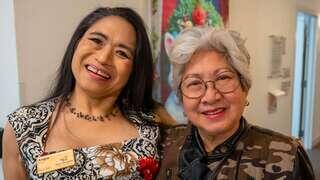 Two smiling women pose together, one dressed in a colorful outfit with a floral design and the other wearing glasses and a chic coat.