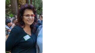 A smiling woman with curly hair and glasses stands in a lively outdoor setting, wearing a black top and a name tag.