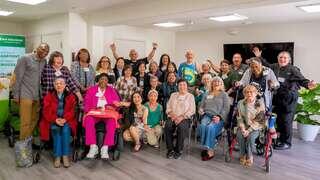 A diverse group of smiling individuals gathers together, celebrating community and connection, with some holding up their hands in joy.