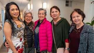 Five women of varying ages stand together, smiling and dressed in colorful outfits, in a well-lit indoor setting.