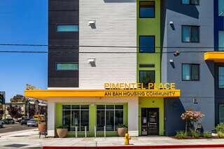 A modern multi-colored building with a bright sign reading "Pimentel Place" stands as part of a housing community, featuring large windows and a welcoming entrance.