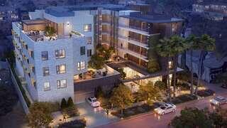 A modern multi-story building features balconies, landscaped gardens, and outdoor seating. Cars are parked along the lit street.