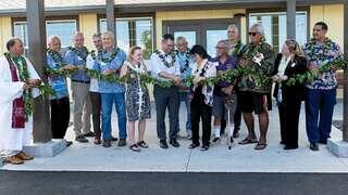 A diverse group of individuals wearing floral leis participates in a ceremonial event, holding greenery together in a celebratory setting.