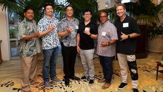 A group of six men poses together in a well-lit setting, all smiling and making a hand gesture. They wear casual and patterned clothing.