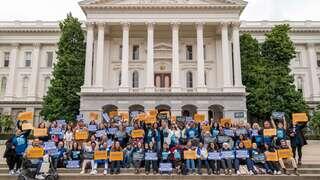 A large group of people gathers in front of a grand building, holding signs advocating for change. Trees frame the background.