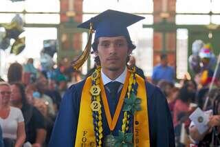 A graduate in a blue cap and gown stands proudly, adorned with multiple achievement cords, amidst a crowd celebrating the occasion.