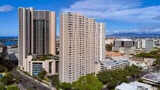 Aerial view of modern high-rise buildings with a clear blue sky, lush greenery, and distant ocean, showcasing a vibrant urban landscape.