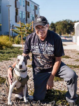 A veteran in a USMC shirt kneels beside his dog in a sunny outdoor setting, surrounded by greenery and modern buildings.