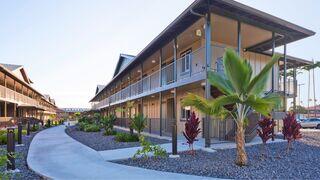 Two-story buildings with balconies surround a landscaped pathway, featuring tropical plants and stones, creating a serene, modern atmosphere.