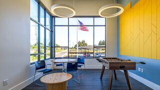 A modern lounge area features large windows with an American flag, a foosball table, and contemporary seating against a bright yellow wall.