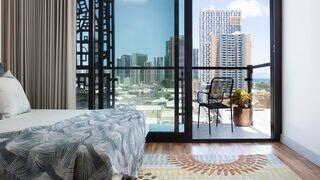 A cozy bedroom with a patterned bedspread, leading to a balcony that offers a view of city skyscrapers and a distant ocean.