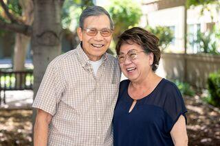 A cheerful elderly couple smiles warmly at each other in a sunlit outdoor setting, showcasing joy and companionship.
