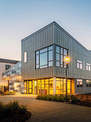 A modern building features sleek, metallic siding and large windows, illuminated softly at dusk, surrounded by landscaping and pathways.