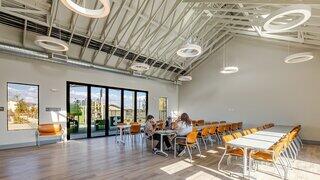 A bright, modern dining space with high ceilings, large windows, and a long table surrounded by chairs, creating a welcoming atmosphere.