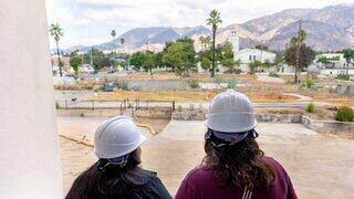 Two individuals in hard hats observe a construction site in Altadena, California with mountains in the background, surrounded by greenery and palm trees.