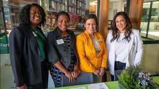 Four women leaders together, smiling and dressed professionally, in a bright, modern space with greenery and a table in front of them.