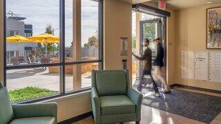 A bright, modern lobby features cozy seating and large windows. Two people walk through the door, leading to an outdoor area with yellow umbrellas.