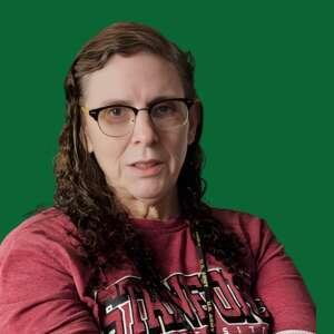 A person with curly hair, wearing glasses and a Stanford sweatshirt, has a thoughtful expression against a green backdrop.