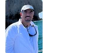 A man in a white shirt and baseball cap stands on a boat with a rocky coastline and ocean in the background, enjoying a sunny day.