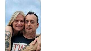 A couple poses closely together, smiling against a cloudy sky. Both have tattoos, showcasing their affectionate connection.