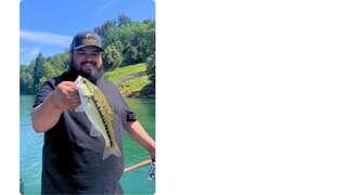A smiling person holds a freshly caught fish while standing on a boat, surrounded by clear water and lush greenery.